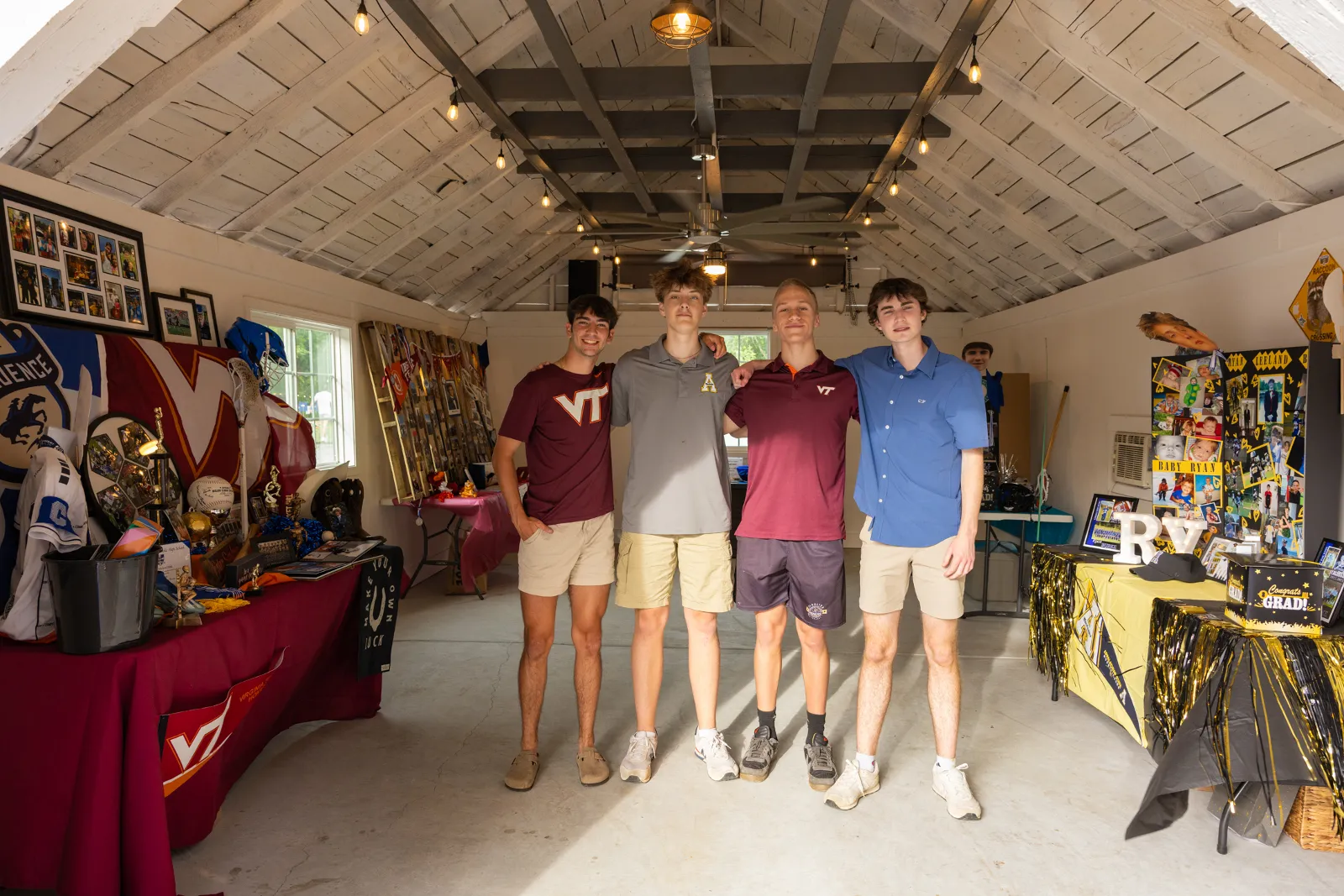 Four young men stand arm in arm in a decorated garage. They are wearing casual shorts and shirts, with one shirt featuring the letters "VT." The garage is adorned with various memorabilia, including sports items and string lights. Huntersville event venue