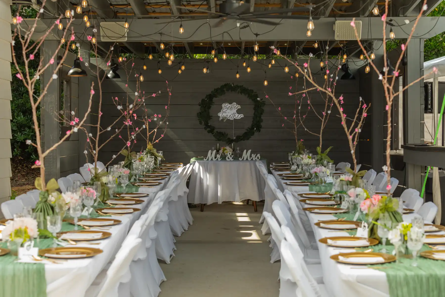 A wedding reception setup at an outdoor event venue features two long tables aligned under a covered patio. Adorned with white linens, floral centerpieces, and elegant place settings, the space gleams with overhead string lights and decorative branches. A "Mr & Mrs" sign adds to the romantic atmosphere. Huntersville event venue