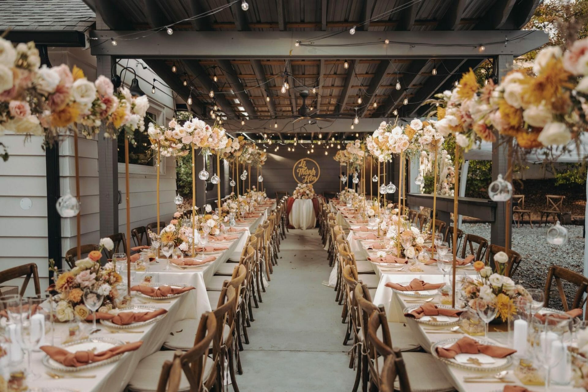 A beautifully decorated outdoor event venue hosts a wedding reception with long banquet tables adorned with floral centerpieces, string lights overhead, and neatly arranged table settings featuring pink napkins. A "Just Married" sign graces the open space event venue in the background. Huntersville event venue