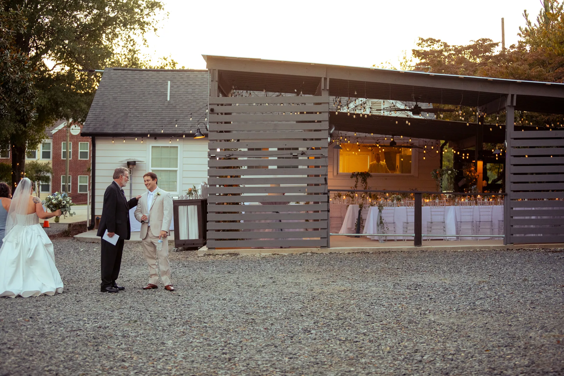 A bride and groom are speaking to a man in a suit outside a rustic, open-air venue decorated with string lights. Tables are set for an event inside the structure, and the sun is setting in the background. Huntersville event venue
