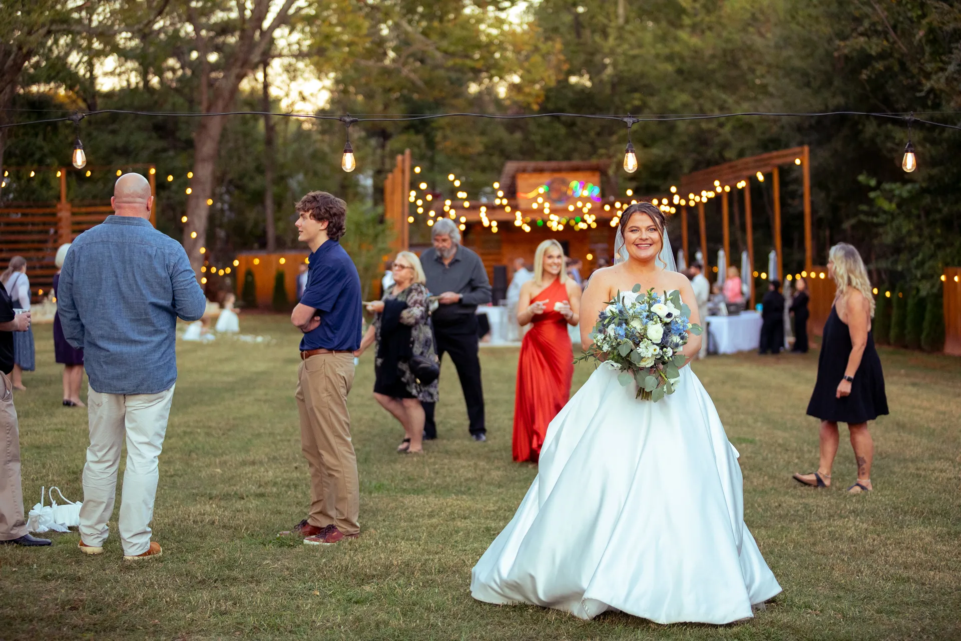 A bride in a white gown holds a bouquet, smiling, while standing outdoors at a wedding reception. Guests in formal attire mingle in the background under string lights. Trees and a decorated archway are visible in the background. Huntersville event venue