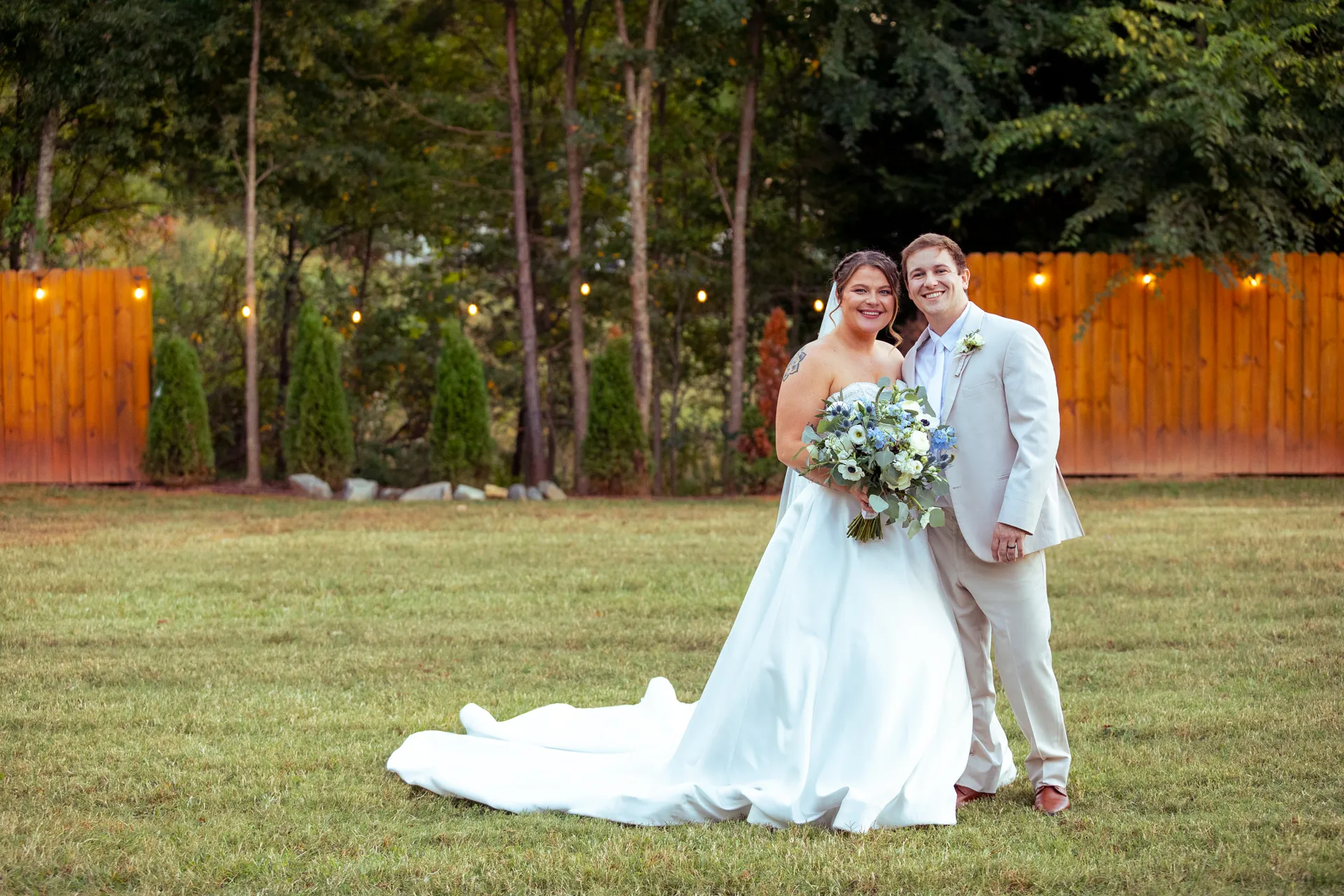 A bride and groom stand together on a grassy field, with the bride in a white gown holding a bouquet and the groom in a beige suit. A wooden fence and trees with string lights are in the background. They are both smiling. Huntersville event venue