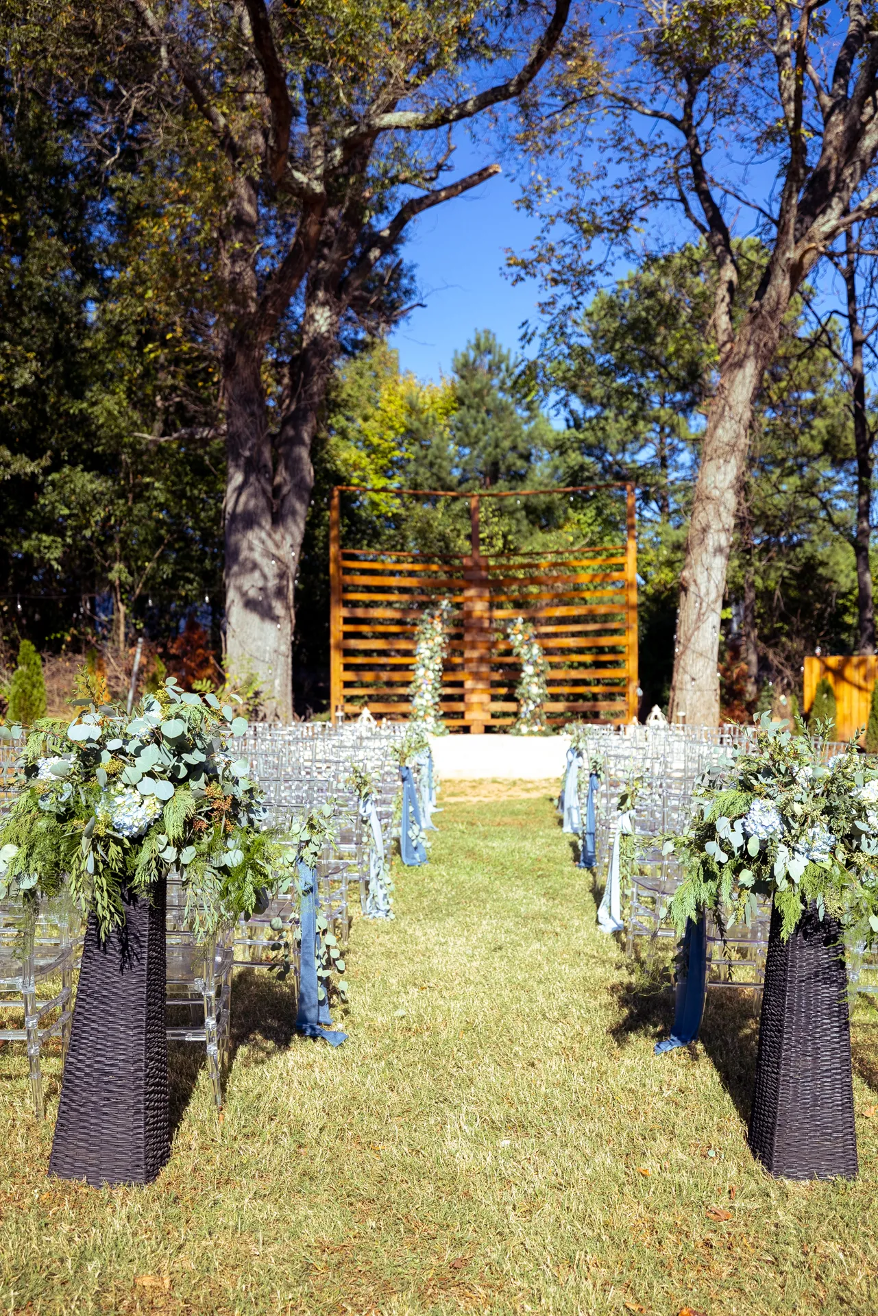 Outdoor wedding ceremony setup with clear chairs lined in rows facing a wooden backdrop adorned with greenery. Two tall flower arrangements flank the aisle on a grassy lawn surrounded by trees under a clear blue sky. Huntersville event venue