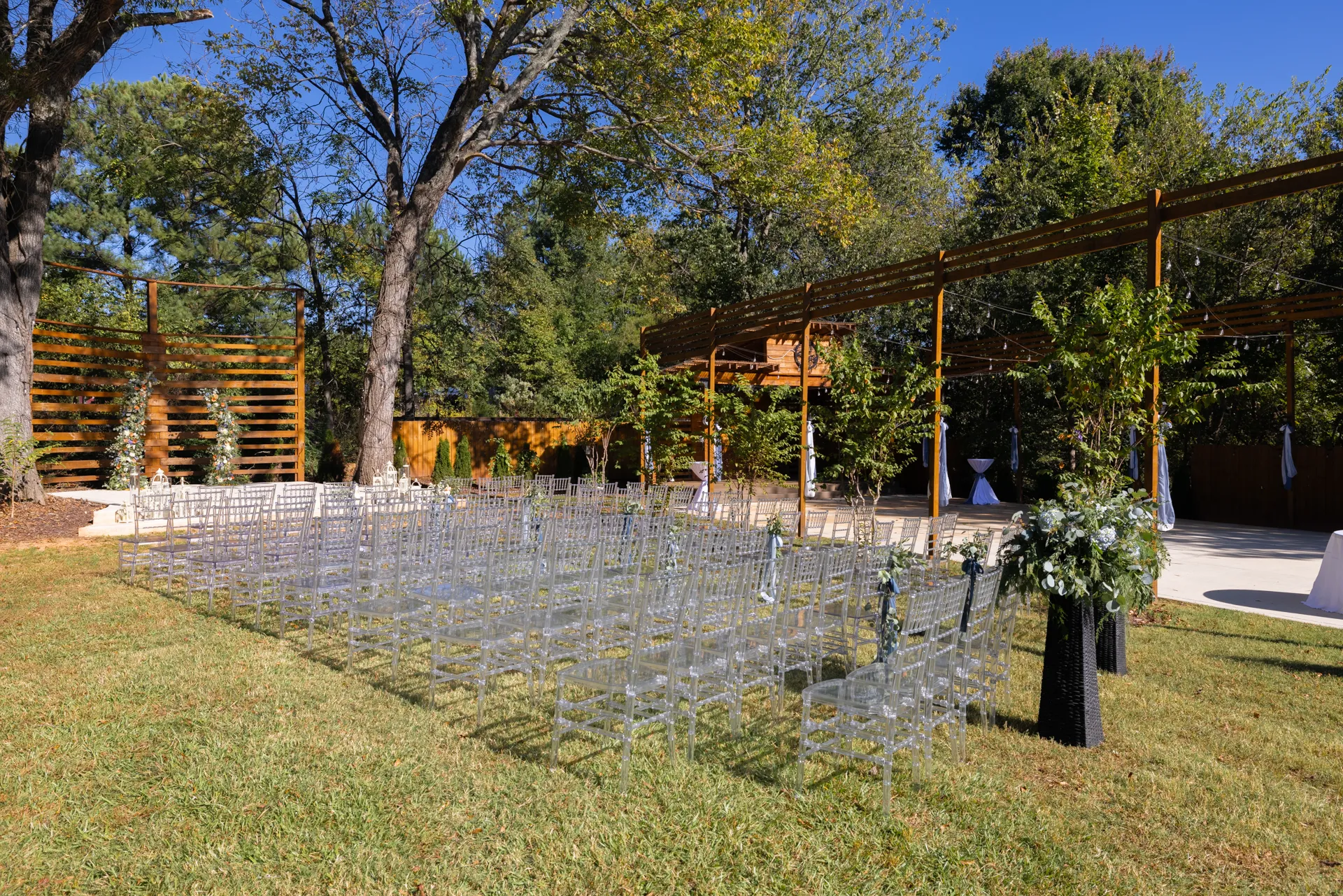An outdoor wedding setup with rows of transparent chairs on grass, facing a wooden pergola decorated with greenery. Tall trees surround the area, creating a natural backdrop under a clear blue sky. Huntersville event venue