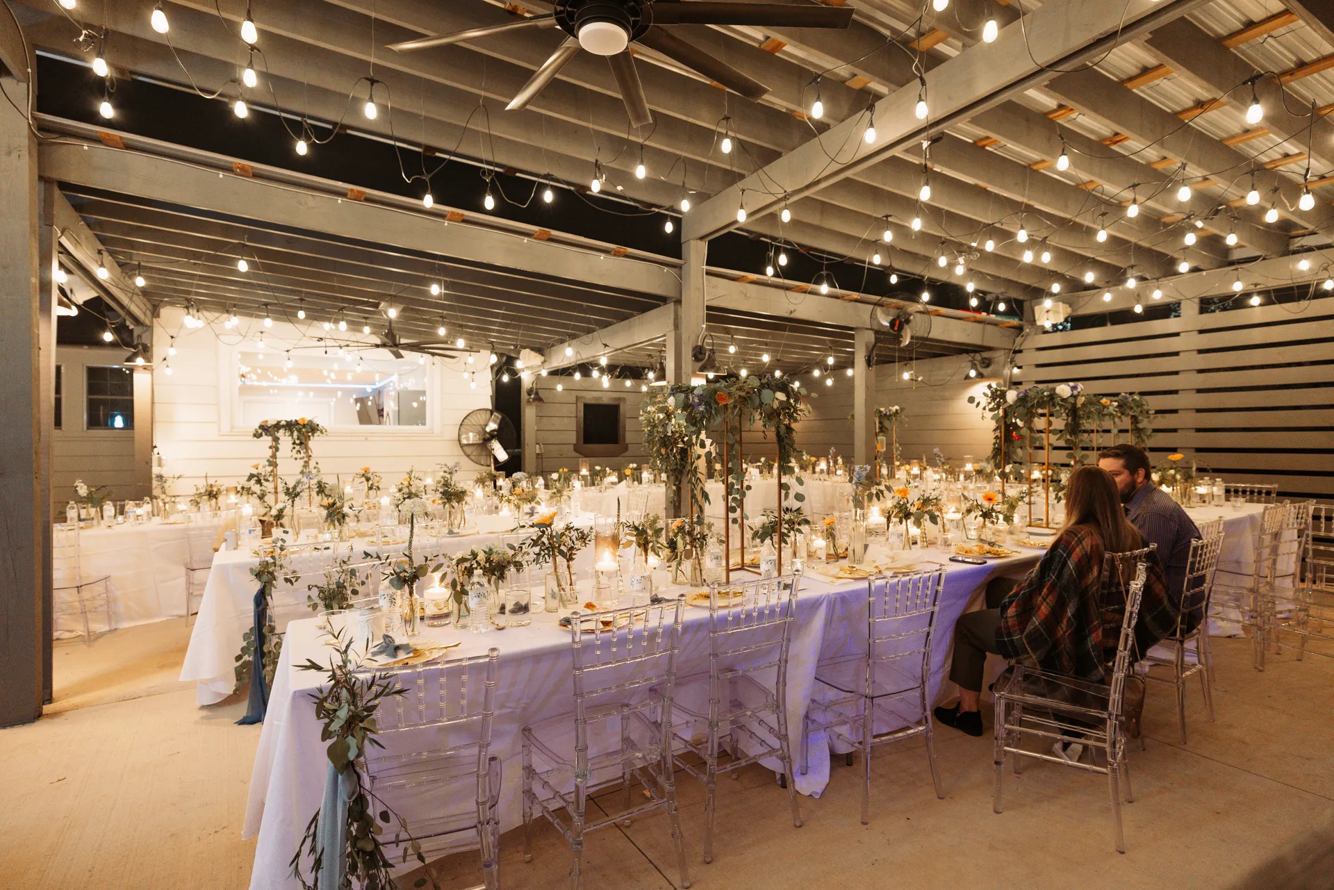 A warmly lit outdoor event space with long tables draped in white tablecloths, decorated with greenery and candles. String lights are hanging from the ceiling, and a couple is seated at the end of one table. Huntersville event venue