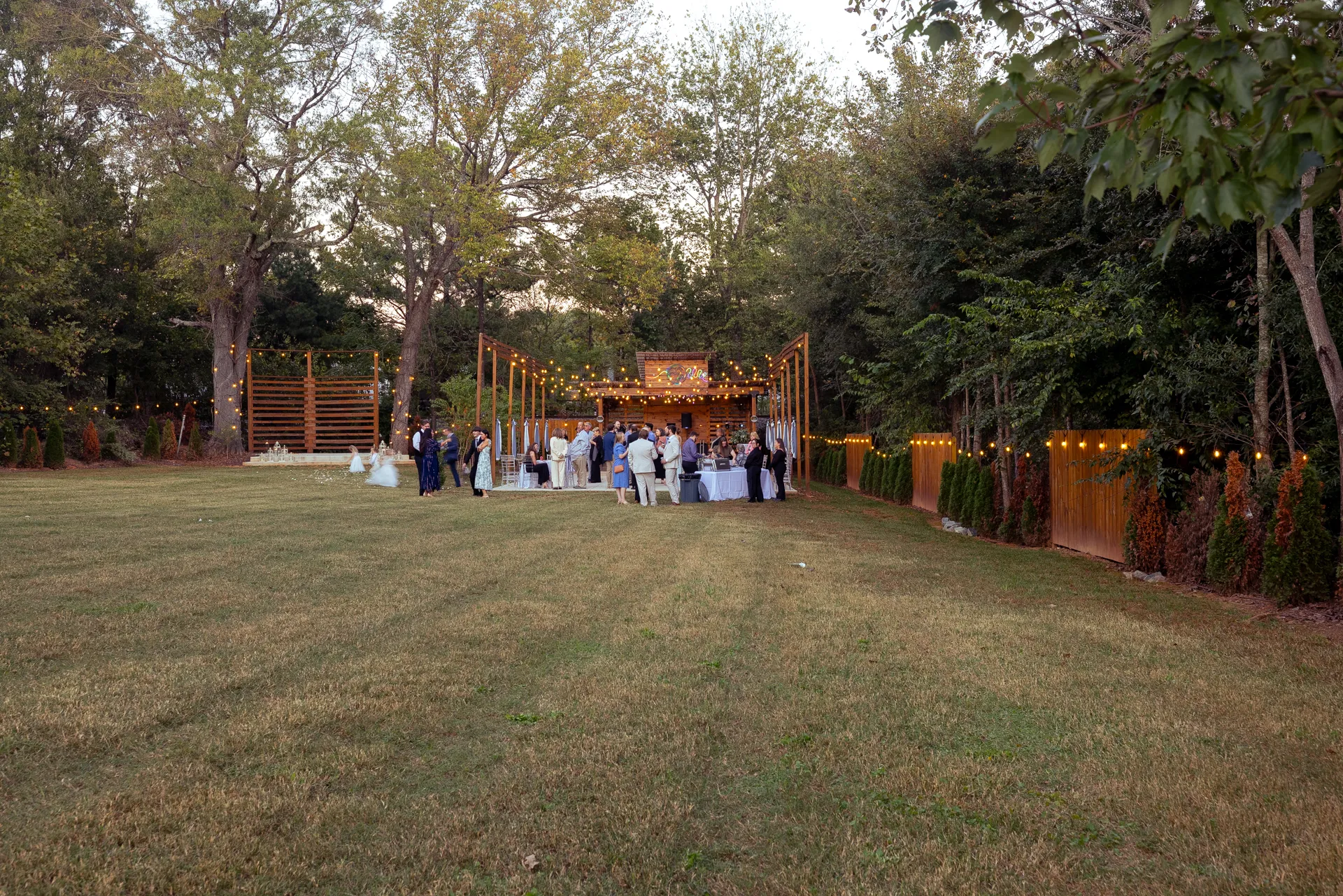 A group of people gathered at an outdoor event in a grassy area surrounded by trees. The setting features a wooden structure with string lights and tables with white tablecloths. The scene is lively and festive. Huntersville event venue