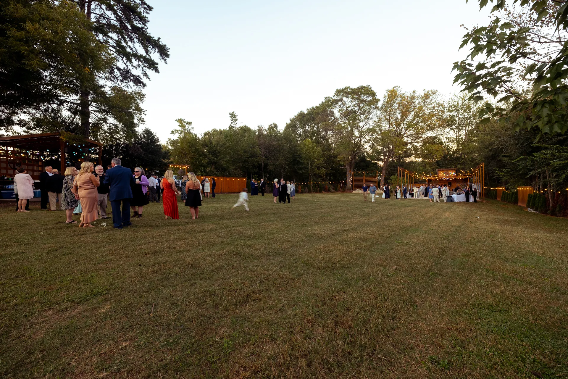 A group of people are gathered outdoors at an event, standing on a large grassy field. Trees surround the area, and string lights are visible. Evening light adds a warm ambiance to the scene. Huntersville event venue