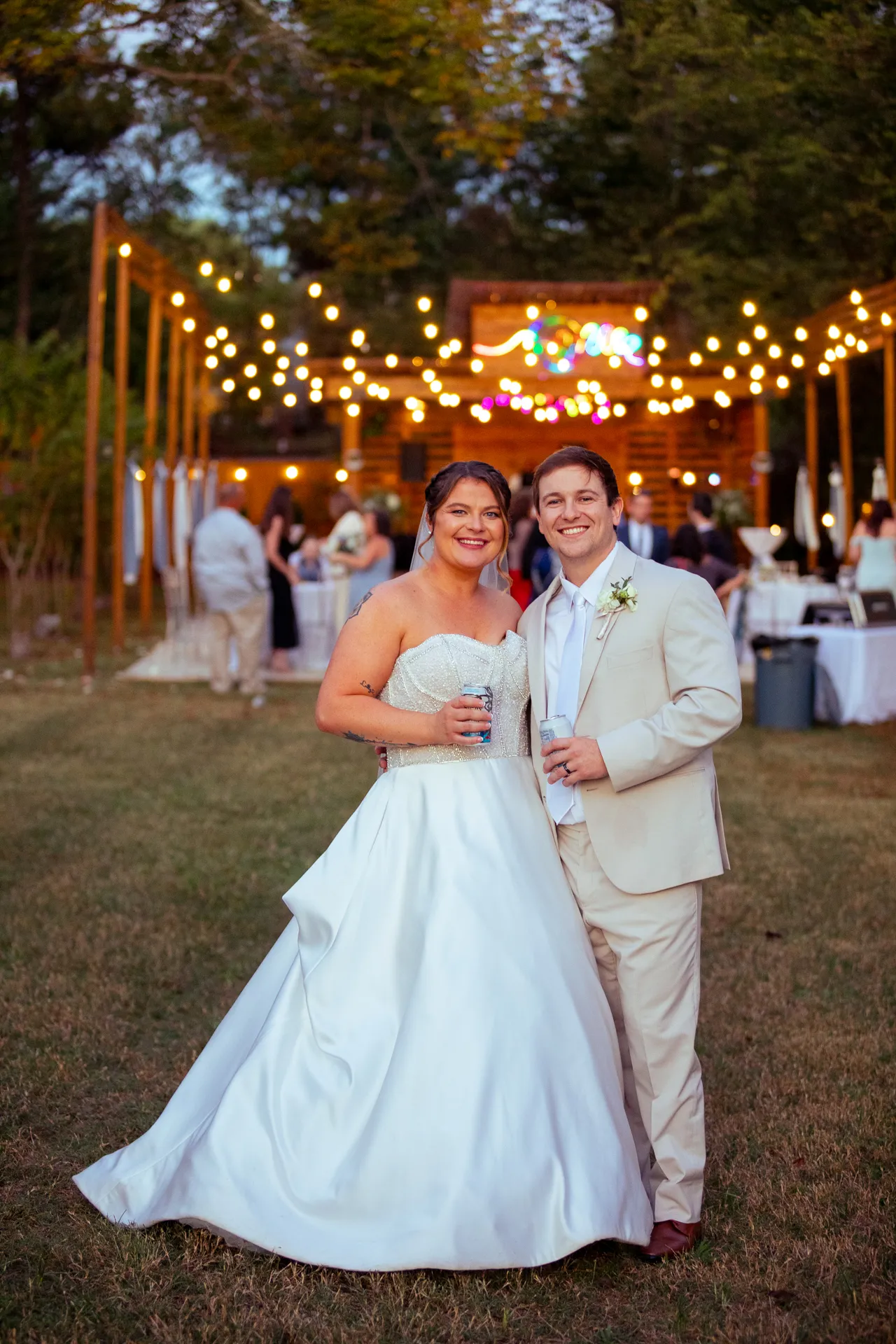 A bride and groom smile at an outdoor wedding reception. The bride wears a white gown, and the groom wears a beige suit. They hold drinks, standing on grass under string lights, with guests and tables in the background. Huntersville event venue