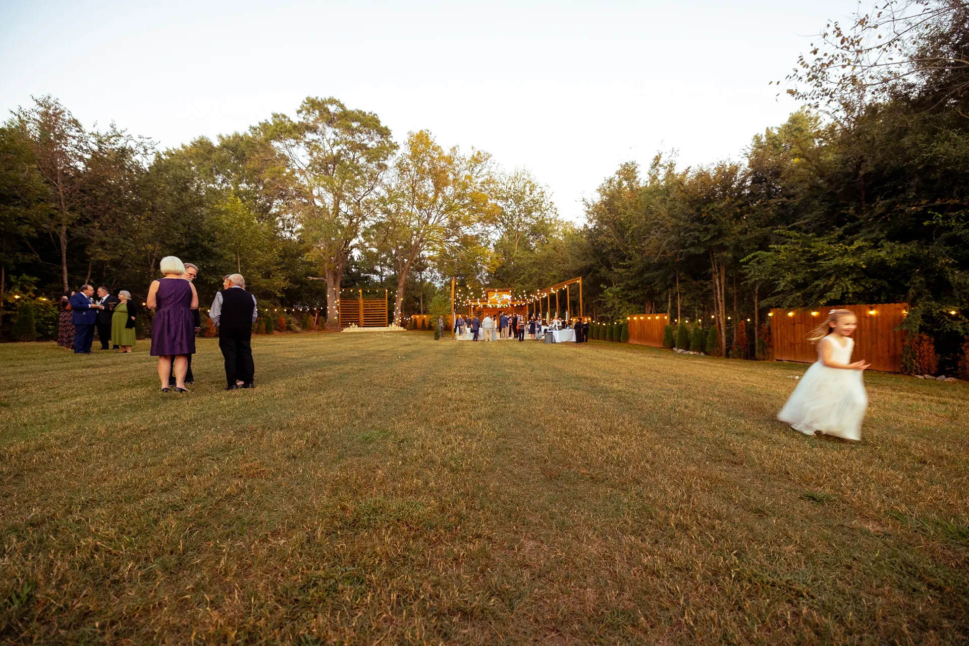 A grassy field with people gathering for an outdoor wedding reception. Guests are dressed in formal attire, and a child in a white dress runs in the foreground. Trees surround the area, and string lights add a warm ambiance. Huntersville event venue