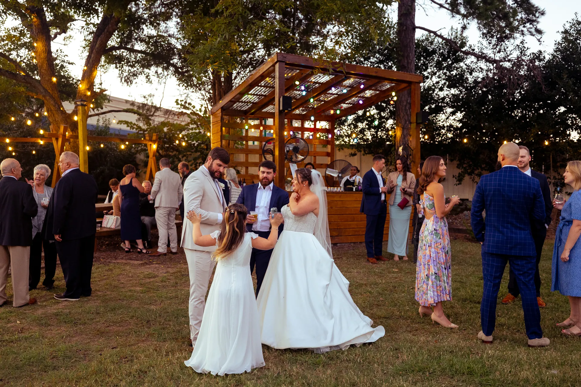 A group of people in formal attire dance on a grassy area at an outdoor wedding reception. String lights hang above, and there is a wooden bar in the background. Trees and guests enjoying the event are also visible. Huntersville event venue