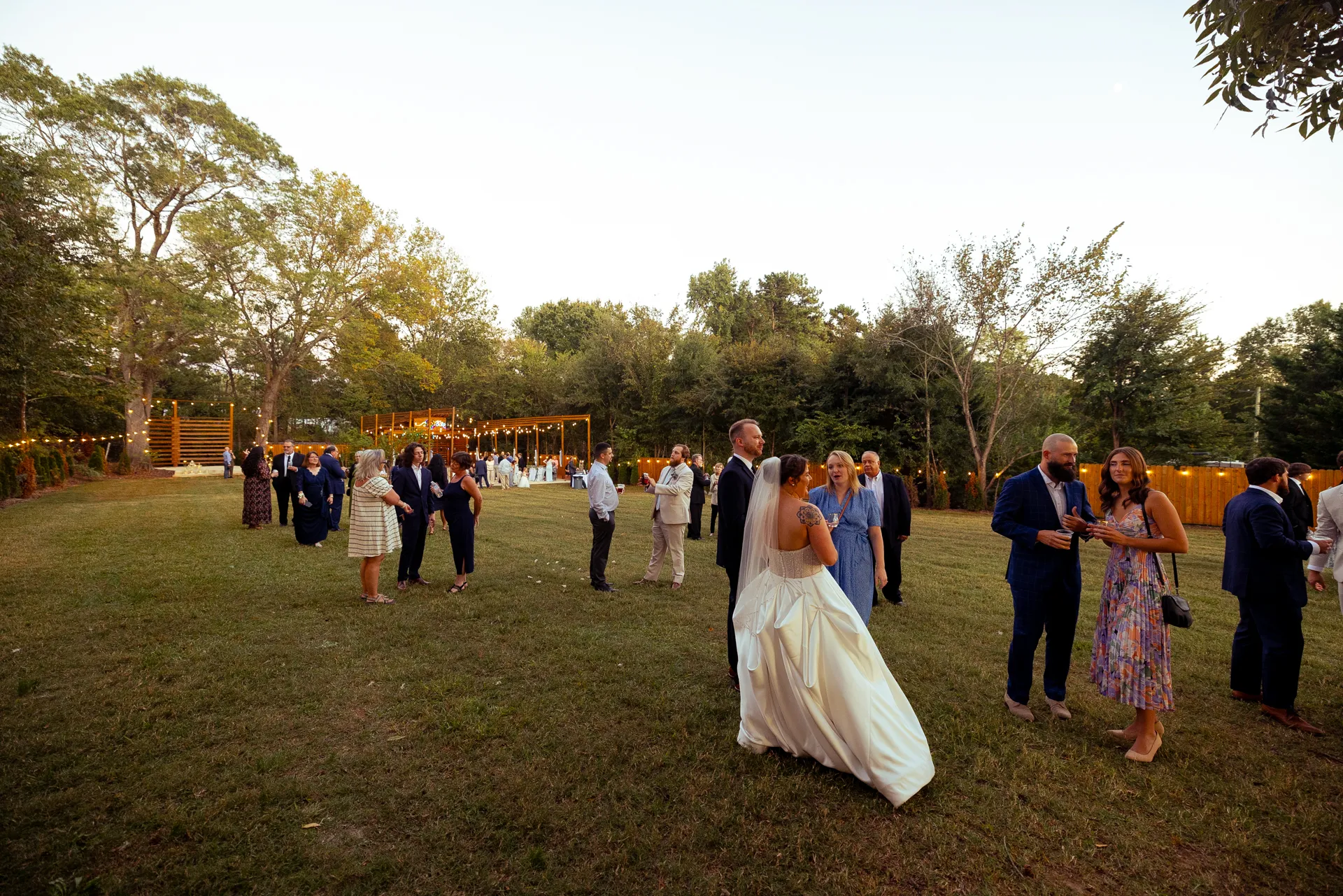 A bride and groom walk through a grassy outdoor venue, surrounded by wedding guests. The scene is set in the evening with decorative string lights in the background, and guests are dressed in formal attire. Trees and a wooden structure are visible. Huntersville event venue